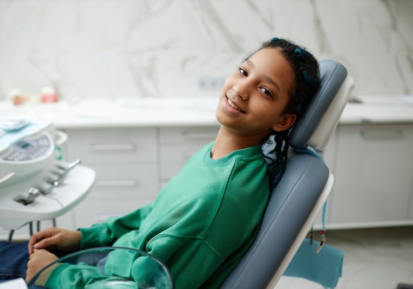 Adolescente nera seduta sulla poltrona del dentista e sorridente alla telecamera durante il controllo