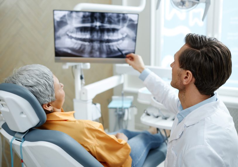 Back view of male dentist pointing at tooth X-ray image on screen during consultation in modern dental clinic